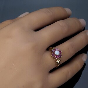 Puede incluir: Un anillo de oro con un dise&ntilde;o floral, con una piedra central blanca y piedras rosas circundantes. El anillo tiene una banda delicada en forma de hoja. El anillo est&aacute; en una mano.