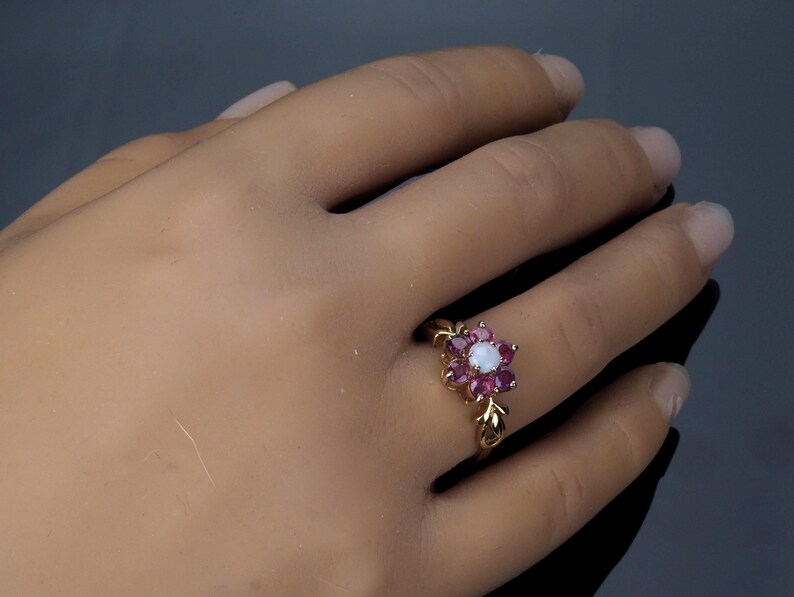 Puede incluir: Un anillo de oro con un dise&ntilde;o floral, con una piedra central blanca y piedras rosas circundantes. El anillo tiene una banda delicada en forma de hoja. El anillo est&aacute; en una mano.