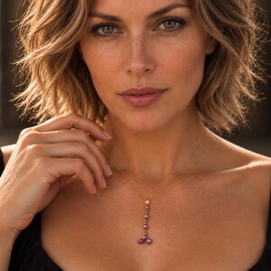 May include: A gold chain necklace featuring a vertical drop pendant composed of five small, round, vibrant pink gemstones arranged in a linear pattern that branches into two at the bottom, resting against a black garment.
