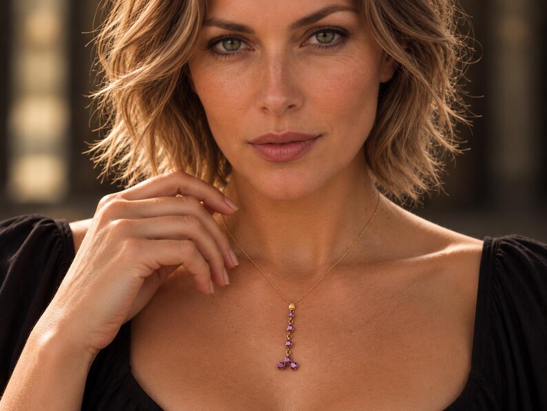 May include: A gold chain necklace featuring a vertical drop pendant composed of five small, round, vibrant pink gemstones arranged in a linear pattern that branches into two at the bottom, resting against a black garment.