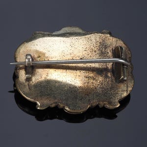 May include: A vintage brooch with a decorative, ornate design. The back of the brooch is visible, showing a pin clasp and a tarnished, gold-toned metal surface. The brooch has an irregular, scalloped edge.