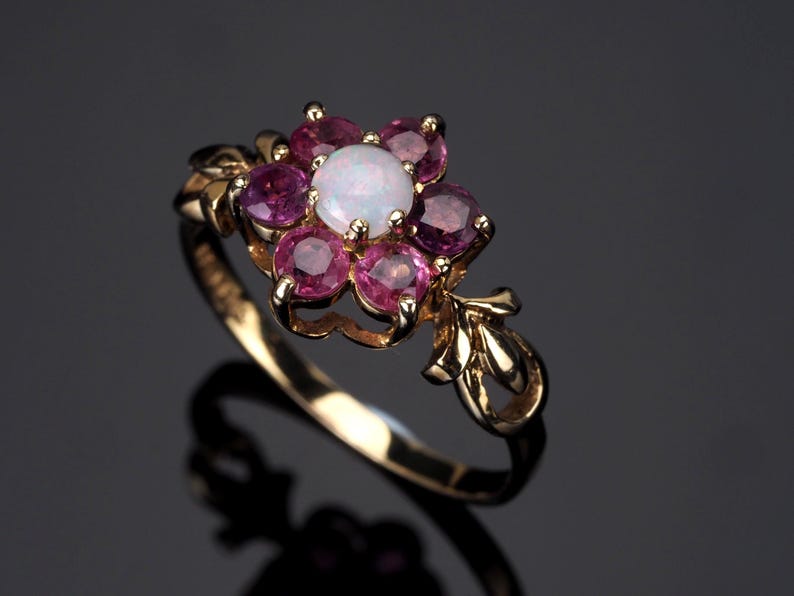 Puede incluir: Un anillo de oro con un dise&ntilde;o floral. El centro de la flor es un &oacute;palo blanco, rodeado de seis piedras preciosas rosas. El anillo tiene una banda delicada de inspiraci&oacute;n floral.