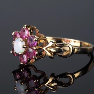 Puede incluir: Un anillo de oro con un dise&ntilde;o floral, con una &oacute;palo blanco central y gemas rosas. La banda del anillo tiene un dise&ntilde;o ornamentado. El anillo se muestra sobre una superficie reflectante.