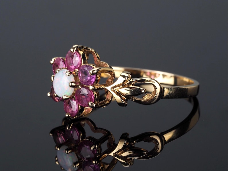 Puede incluir: Un anillo de oro con un dise&ntilde;o floral, con una &oacute;palo blanco central y gemas rosas. La banda del anillo tiene un dise&ntilde;o ornamentado. El anillo se muestra sobre una superficie reflectante.