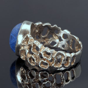 May include: A silver ring with a blue gemstone set in a textured band. The band is made of a series of interconnected loops, creating a unique and intricate design.
