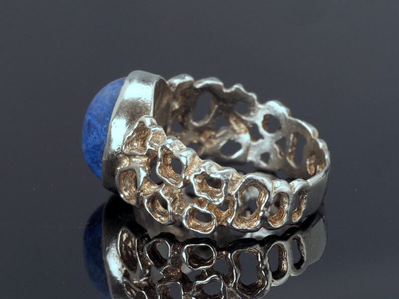 May include: A silver ring with a blue gemstone set in a textured band. The band is made of a series of interconnected loops, creating a unique and intricate design.