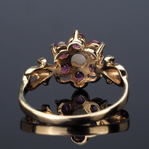 Puede incluir: Un anillo de oro con un dise&ntilde;o floral, con una perla central y gemas rosas circundantes. La banda del anillo es lisa y el engaste tiene detalles intrincados. El anillo se muestra sobre una superficie reflectante.