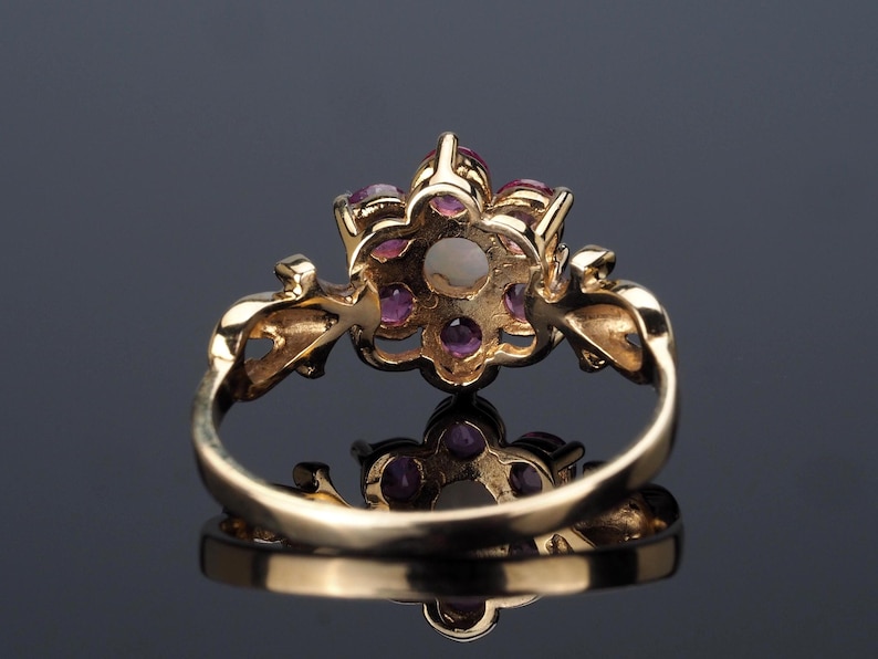 Puede incluir: Un anillo de oro con un dise&ntilde;o floral, con una perla central y gemas rosas circundantes. La banda del anillo es lisa y el engaste tiene detalles intrincados. El anillo se muestra sobre una superficie reflectante.
