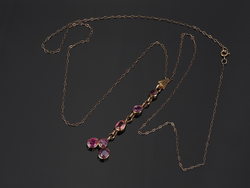 May include: A gold chain necklace featuring a vertical drop pendant set with six oval-shaped pink gemstones. The pendant consists of a single line of four stones leading to a base with two additional stones arranged in a cluster at the bottom.