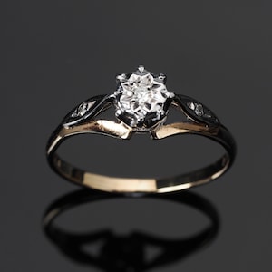 May include: A gold and silver ring with a single, round diamond in the center. The diamond is set in a six-prong setting and is surrounded by smaller diamonds.