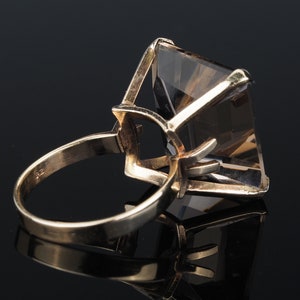 May include: A gold ring with a large, clear, faceted gemstone. The gemstone is set in a prong setting and has a square shape.