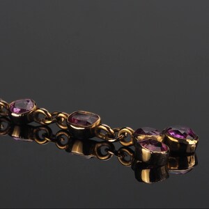 May include: A gold-toned chain bracelet featuring a series of bezel-set, faceted purple gemstones. The links are connected in a linear fashion, with a cluster of three stones at the end, all reflecting against a dark, smooth surface.