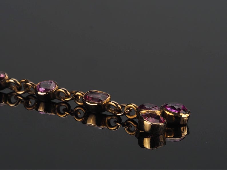 May include: A gold-toned chain bracelet featuring a series of bezel-set, faceted purple gemstones. The links are connected in a linear fashion, with a cluster of three stones at the end, all reflecting against a dark, smooth surface.