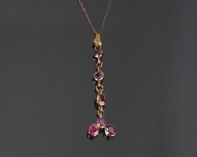 May include: A gold pendant necklace featuring a vertical drop design with four individual pink gemstones set in gold, ending in a cluster of three additional pink gemstones at the base, suspended from a delicate gold chain.