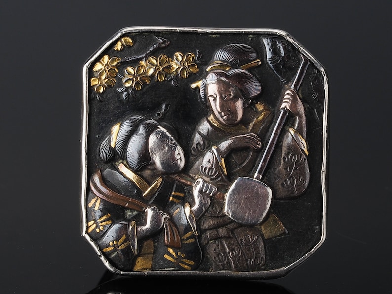 Pu&ograve; includere: Un netsuke giapponese in argento e oro del periodo Edo che raffigura due donne, una delle quali suona uno shamisen, uno strumento musicale a tre corde. Il netsuke &egrave; finemente intagliato con dettagli dei loro vestiti e dello strumento.