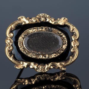 May include: An antique gold-toned brooch with a black enamel background. The brooch features an oval center with a clear cover, surrounded by a gold frame with ornate detailing. The pin is a classic piece of jewelry.