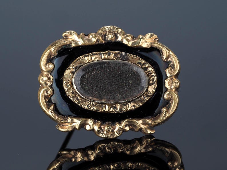 May include: An antique gold-toned brooch with a black enamel background. The brooch features an oval center with a clear cover, surrounded by a gold frame with ornate detailing. The pin is a classic piece of jewelry.