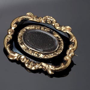 May include: An antique gold-toned brooch with an oval, dark-colored center stone. The brooch features an ornate, swirling design around the central stone and a pin on the back. The brooch is set against a dark background.