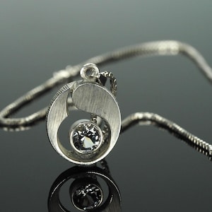 May include: A silver pendant necklace with a circular design and a clear gemstone at the center. The pendant hangs from a delicate chain. The necklace is displayed on a reflective surface.