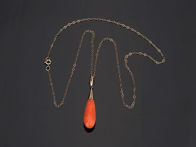 Pu&ograve; includere: Una collana a catena in oro con un ciondolo a forma di goccia realizzato in corallo arancione liscio e vibrante. Il ciondolo &egrave; fissato alla catena tramite una contromaglia in oro lucido e la collana include una piccola chiusura a molla.