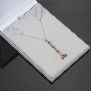 May include: A gold chain necklace featuring a vertical drop pendant adorned with multiple round, faceted pink gemstones. The necklace is displayed inside a light gray presentation box with a white interior lining and a hinged lid.
