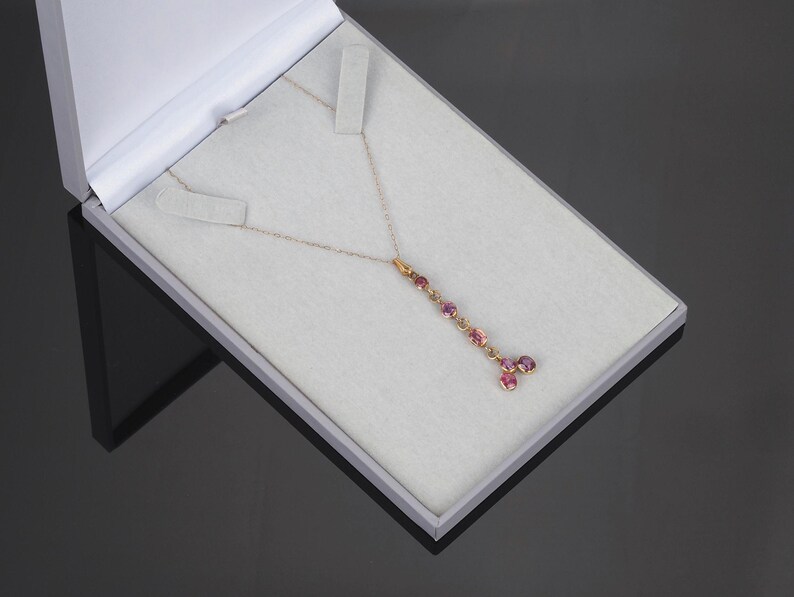 May include: A gold chain necklace featuring a vertical drop pendant adorned with multiple round, faceted pink gemstones. The necklace is displayed inside a light gray presentation box with a white interior lining and a hinged lid.