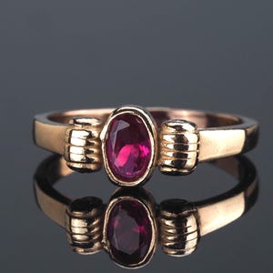 Può includere: Un anello d'oro con una grande gemma di rubino ovale. L'anello ha una fascia semplice con dettagli a coste su entrambi i lati del rubino. L'anello è esposto su una superficie riflettente, creando un effetto specchio.