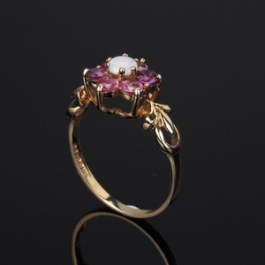 Puede incluir: Un anillo de oro con un dise&ntilde;o floral, con una piedra central blanca y piedras rosas circundantes. El anillo tiene una banda ornamentada con detalles de hojas. El anillo est&aacute; sobre un fondo oscuro.
