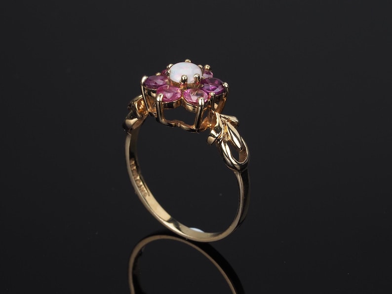 Puede incluir: Un anillo de oro con un dise&ntilde;o floral, con una piedra central blanca y piedras rosas circundantes. El anillo tiene una banda ornamentada con detalles de hojas. El anillo est&aacute; sobre un fondo oscuro.