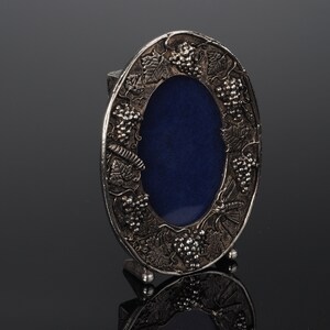 May include: Silver-toned oval picture frame with a blue velvet backing and an ornate design of grapes and leaves around the edge.