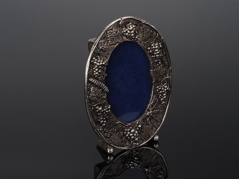 May include: Silver-toned oval picture frame with a blue velvet backing and an ornate design of grapes and leaves around the edge.