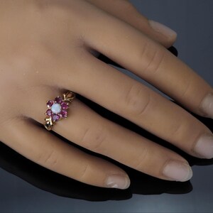 Puede incluir: Un anillo de oro con un dise&ntilde;o floral, con una piedra blanca central rodeada de piedras rojas oscuras m&aacute;s peque&ntilde;as. El anillo se muestra en una mano, destacando sus intrincados detalles y contraste de color. El dise&ntilde;o del anillo evoca un arreglo floral.