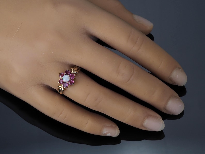 Puede incluir: Un anillo de oro con un dise&ntilde;o floral, con una piedra blanca central rodeada de piedras rojas oscuras m&aacute;s peque&ntilde;as. El anillo se muestra en una mano, destacando sus intrincados detalles y contraste de color. El dise&ntilde;o del anillo evoca un arreglo floral.
