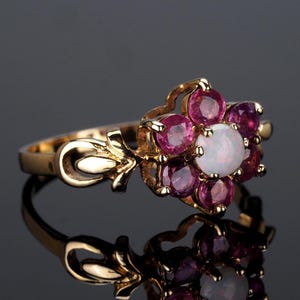 Puede incluir: Un anillo de oro con un dise&ntilde;o floral. El anillo presenta una &oacute;palo blanco central, rodeado de seis piedras preciosas redondas de color rosa oscuro m&aacute;s peque&ntilde;as. La banda tiene detalles decorativos en forma de hoja.
