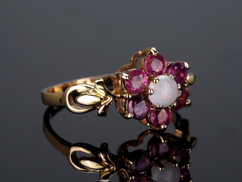 Puede incluir: Un anillo de oro con un dise&ntilde;o floral. El anillo presenta una &oacute;palo blanco central, rodeado de seis piedras preciosas redondas de color rosa oscuro m&aacute;s peque&ntilde;as. La banda tiene detalles decorativos en forma de hoja.