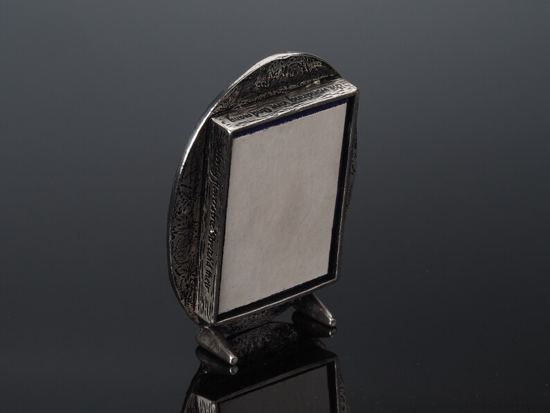 May include: A silver-toned oval picture frame with a textured surface and a small stand. The frame has a blue and white border and the text "The Pleasant Street Shop" is engraved on the side.