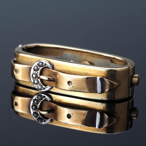 May include: A gold-toned hinged bracelet with a buckle design. The bracelet features two parallel bands with silver-colored buckles adorned with small, dark-colored embellishments. The design is reflected in the dark surface below.