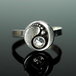 May include: A silver ring with a yin-yang design. The ring features a clear, round gemstone at the center of the yin-yang symbol. The ring has a textured surface and is set against a dark background.