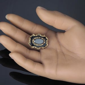 May include: An antique brooch with an oval, light blue center stone, framed by a black enamel border and ornate gold filigree. The brooch is a vintage piece, likely from the Victorian era, and is a small, elegant accessory.