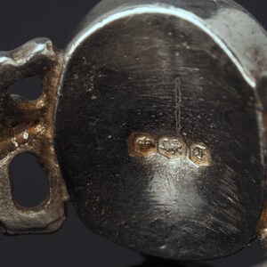 May include: Close-up of the back of a silver ring with a detailed design. The ring has three hallmarks: a crown, a lion, and a letter 'T'.