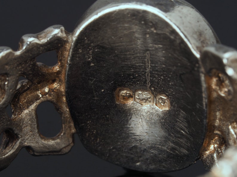May include: Close-up of the back of a silver ring with a detailed design. The ring has three hallmarks: a crown, a lion, and a letter 'T'.