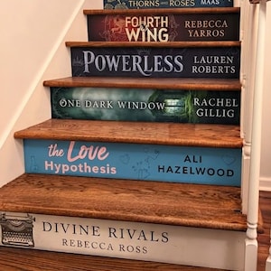 May include: A wooden staircase with risers decorated as book spines, featuring titles including A Court of Thorns and Roses, Fourth Wing, Powerless, One Dark Window, The Love Hypothesis, and Divine Rivals, with authors Sarah J. Maas, Rebecca Yarros, Lauren Roberts, Rachel Gillig, Ali Hazelwood, and Rebecca Ross.