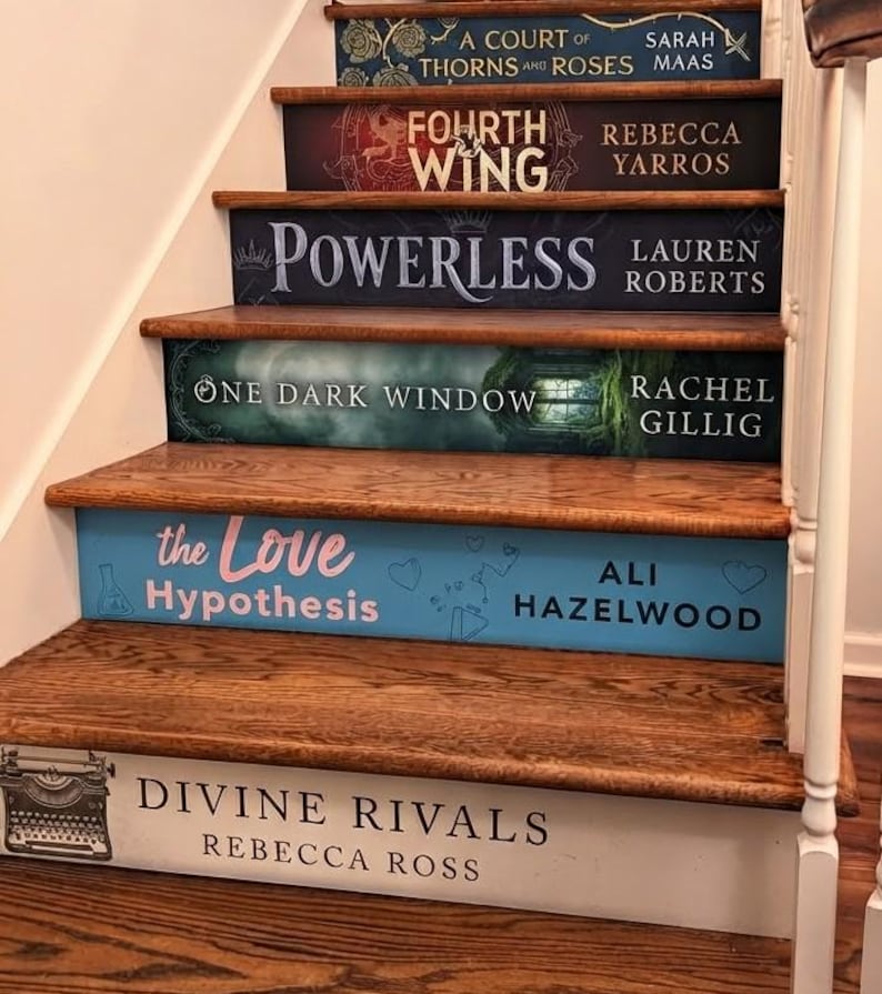 May include: A wooden staircase with risers decorated as book spines, featuring titles including A Court of Thorns and Roses, Fourth Wing, Powerless, One Dark Window, The Love Hypothesis, and Divine Rivals, with authors Sarah J. Maas, Rebecca Yarros, Lauren Roberts, Rachel Gillig, Ali Hazelwood, and Rebecca Ross.