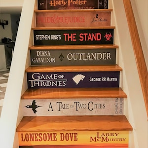 May include: A staircase with risers decorated to resemble book spines, featuring titles such as The Going to Bed Book, Harry Potter, Pride and Prejudice, The Stand, Outlander, Game of Thrones, A Tale of Two Cities, Lonesome Dove, and To Kill a Mockingbird.