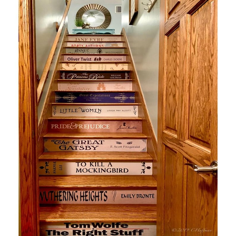 May include: A wooden staircase with risers decorated to resemble book spines, featuring titles such as Jane Eyre, Oliver Twist, The Princess Bride, Little Women, Pride and Prejudice, The Great Gatsby, To Kill a Mockingbird, and Wuthering Heights.