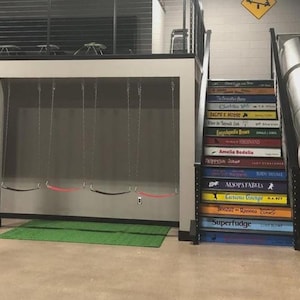 May include: An indoor play area featuring a set of four swings hanging from a loft structure, a staircase with steps painted to resemble book spines with titles like Superfudge and Charlotte's Web, and a metal tube slide next to the stairs.