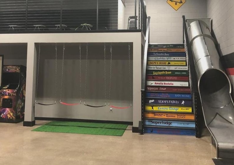 May include: An indoor play area featuring a set of four swings hanging from a loft structure, a staircase with steps painted to resemble book spines with titles like Superfudge and Charlotte's Web, and a metal tube slide next to the stairs.