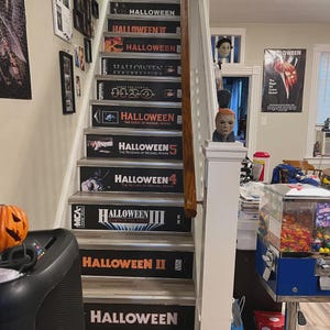 May include: A staircase with risers decorated to resemble VHS tape covers for the Halloween film series, including titles like Halloween, Halloween II, Halloween III, Halloween 4, Halloween 5, H20, and Halloween Resurrection.