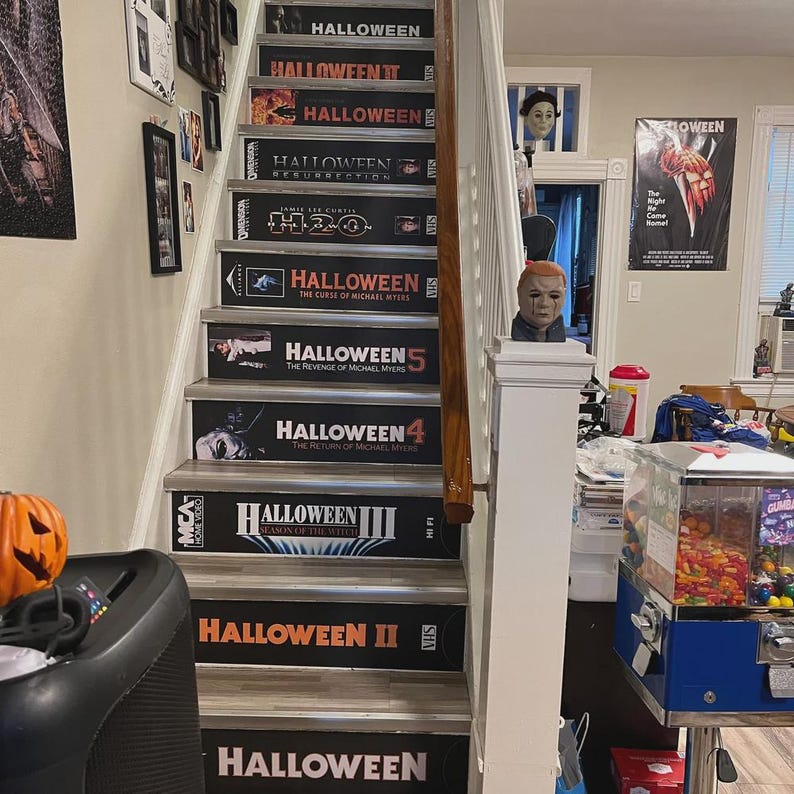 May include: A staircase with risers decorated to resemble VHS tape covers for the Halloween film series, including titles like Halloween, Halloween II, Halloween III, Halloween 4, Halloween 5, H20, and Halloween Resurrection.