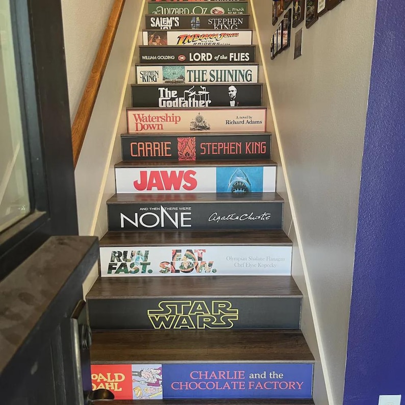 May include: A staircase with risers decorated to resemble book spines, featuring titles such as The Wizard of Oz, Salem's Lot, Indiana Jones, Lord of the Flies, The Shining, The Godfather, Watership Down, Carrie, Jaws, And Then There Were None, Run Fast. Eat Slow., Star Wars, and Charlie and the Chocolate Factory.